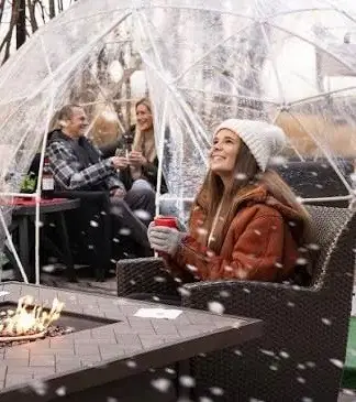 Retreaters warming by a fire table inside a clear-dome igloo as snow falls